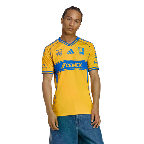 A man models a yellow and blue Tigres UANL soccer jersey with denim shorts against a white background.