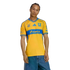 A man models a yellow and blue Tigres UANL soccer jersey with denim shorts against a white background.