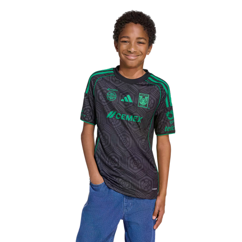 A smiling boy wearing a black and green Tigres UANL soccer jersey and blue jeans stands against a black background.