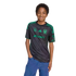 A smiling boy wearing a black and green Tigres UANL soccer jersey and blue jeans stands against a black background.