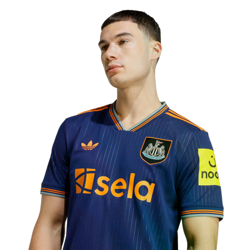 A young man wearing a navy blue Newcastle United football jersey with orange trim and "sela" sponsor logo, set against a black background.