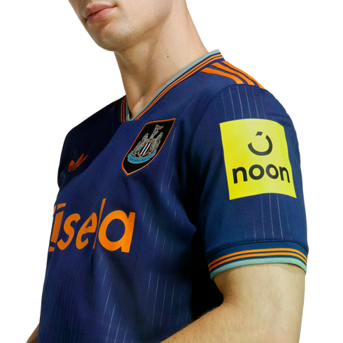 A person wears a blue Newcastle United football jersey with orange accents, featuring the club crest, the "Sela" sponsor on the front, and the "noon" logo on the sleeve.