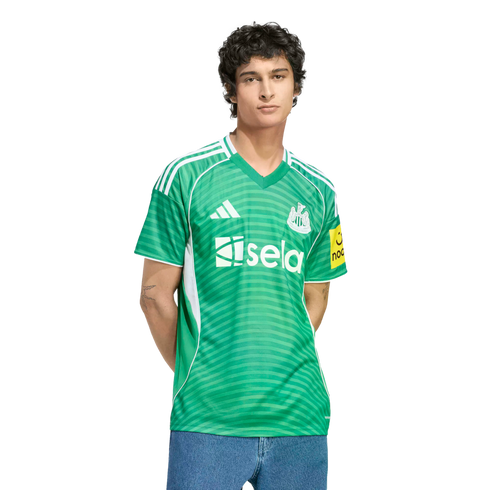 A man wearing a green Newcastle United football jersey with white details and a "sela" sponsor logo, paired with blue jeans against a black background.