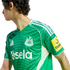 Person in a green Newcastle United football jersey with white Adidas stripes and Sela sponsor logo, shown in profile against a black background.