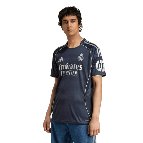 A young man wearing a navy blue Real Madrid soccer jersey with yellow trim and blue jeans, standing against a plain white background.