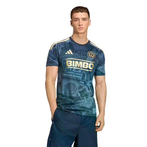 A male model wears a navy Philadelphia Union soccer jersey featuring gold logos and historical graphics, paired with navy shorts against a black backdrop.