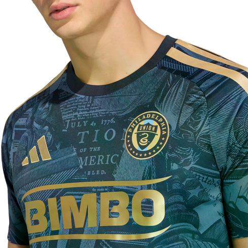 Close-up of a Philadelphia Union soccer jersey with a gold "BIMBO" logo, team crest, and a teal background patterned with historical text.