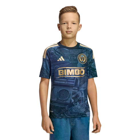 A youth stands wearing a navy blue and gold Philadelphia Union soccer jersey and blue jeans against a solid black background.