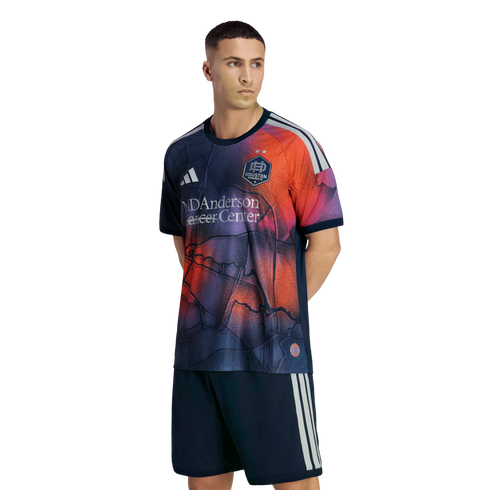 Man modeling a Houston Dynamo FC soccer kit with a navy, orange, and purple abstract patterned jersey and matching navy shorts, against a black background.
