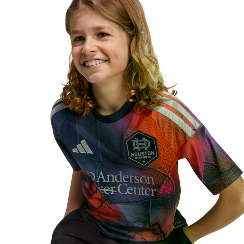 A smiling child with long, wavy light brown hair wears a colorful Houston Dynamo FC soccer jersey against a solid black background.