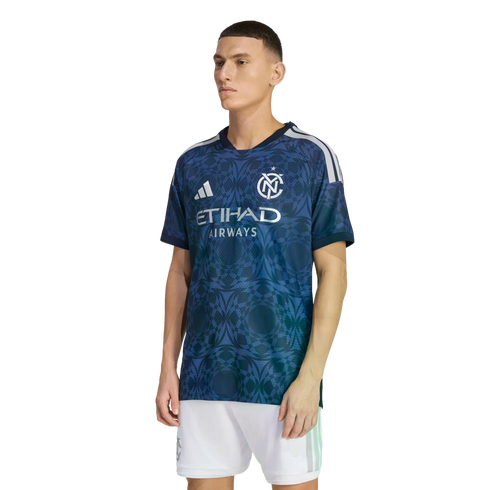 A man models a navy blue NYCFC soccer jersey with a geometric pattern, Adidas logo, and Etihad sponsor, paired with white shorts against a black background.