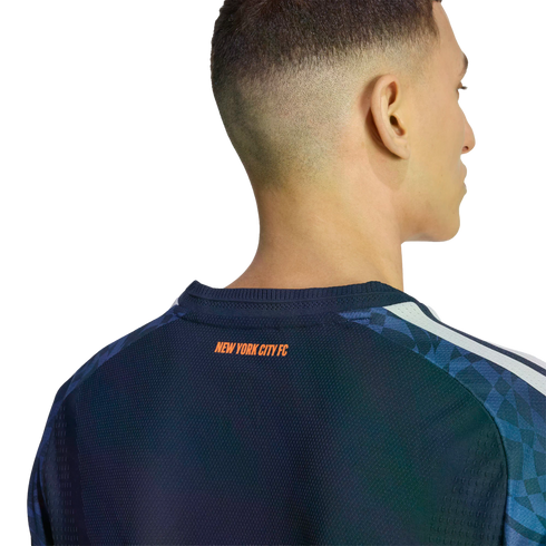 Back view of a man wearing a navy New York City FC soccer jersey with orange text below the collar. The shoulders have a blue geometric pattern and white stripes.