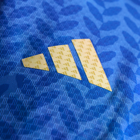 A close-up of a gold three-bar Adidas logo with a repeating pattern, set against blue textured fabric with a leafy design.