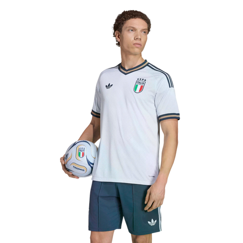A man poses in a white Italy soccer jersey and blue shorts, holding a football against a black background.