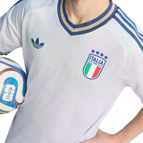 Close-up of a person in a light blue Adidas Italy football jersey with the FIGC crest, holding a FIFA soccer ball against a black background.