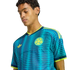 Young man wearing a blue and yellow Colombia soccer jersey with a wavy pattern, looking down against a black background.