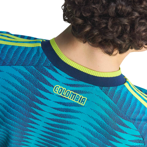 Back view of a person with curly hair wearing a teal patterned soccer jersey with yellow "COLOMBIA" text and lime green accents.