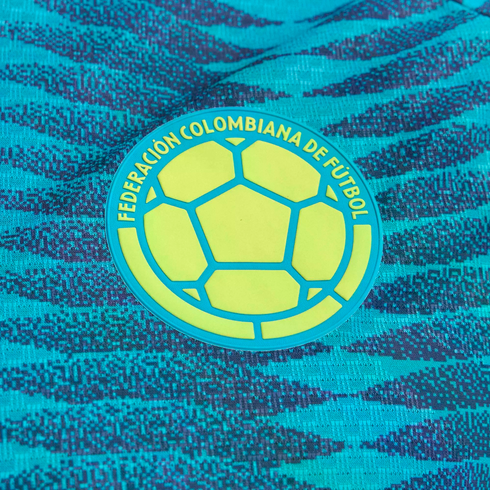 Close-up of the yellow and teal Federación Colombiana de Fútbol badge on a patterned teal soccer jersey.