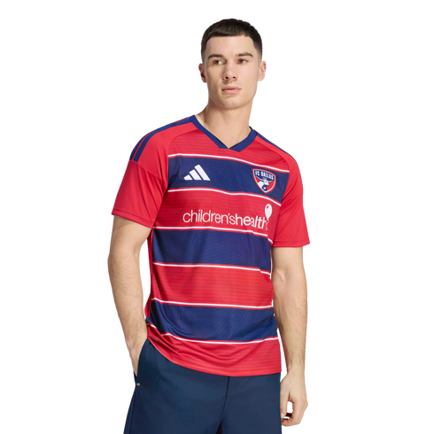 A man modeling a red and blue striped FC Dallas soccer jersey with a white sponsor logo against a black background.