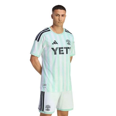 A man models a light green and white striped Austin FC soccer kit with black Adidas details and a YETI logo. He wears matching white shorts against a black background.