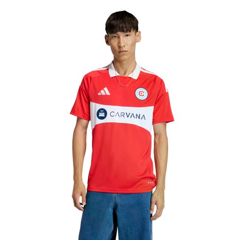 A man models a red Chicago Fire soccer jersey featuring a white chest band with the "CARVANA" logo, paired with blue jeans against a black background.