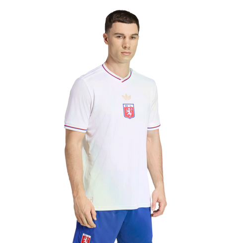 A man models a white Olympique Lyonnais soccer jersey with a gold trefoil logo and blue and red trim, paired with blue shorts, against a solid black background.