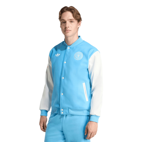 A man models a light blue and white Adidas varsity jacket with an Inter Miami crest and matching light blue pants against a black background.
