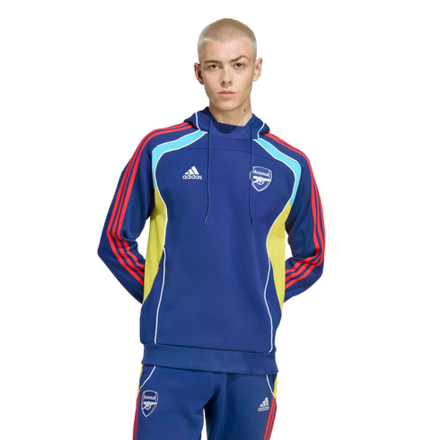 A man with buzzed blonde hair wearing a blue Adidas Arsenal FC tracksuit with red stripes and yellow/light blue accents, posed against a black background.