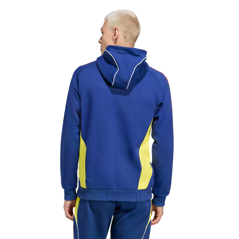 Rear view of a man with blond hair wearing a blue hoodie and matching pants with yellow side panels and white piping, set against a black background.