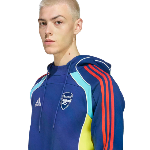 A person with short blonde hair wears a blue Adidas Arsenal FC hoodie with red stripes and yellow and light blue accents against a black background.