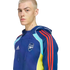 A person with short blonde hair wears a blue Adidas Arsenal FC hoodie with red stripes and yellow and light blue accents against a black background.