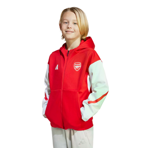 A young blonde boy smiles, wearing a red and white Arsenal zip-up hoodie against a black background.