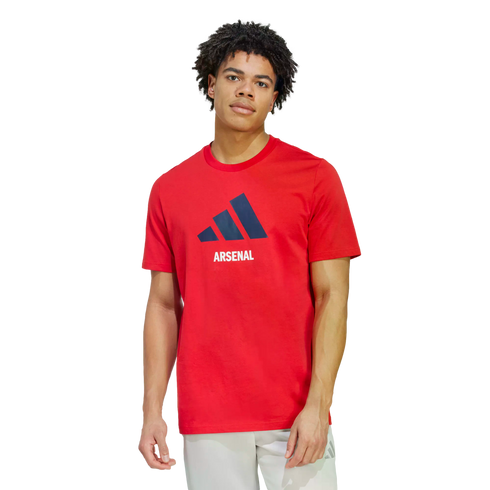 A man wears a red Adidas Arsenal t-shirt with a navy logo and white "ARSENAL" lettering. He is paired with light grey shorts against a solid black background.
