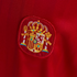 Close-up of the Spanish coat of arms embroidered on a red textured jersey, featuring a gold crown, central shield, and side pillars.