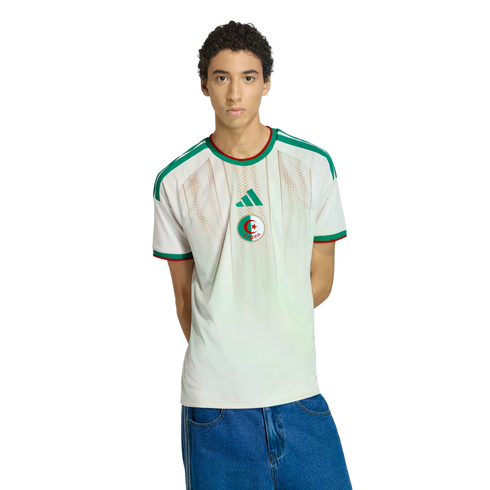 A medium shot of a young man with curly hair wearing an off-white Algeria national team jersey and blue jeans, posed against a black background.