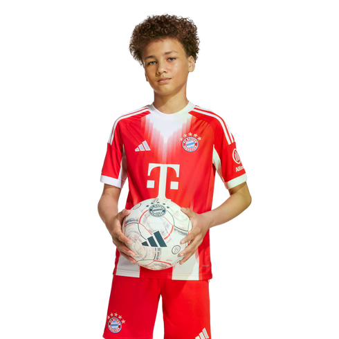 A young boy with curly hair wears a red and white FC Bayern Munich soccer uniform while holding a soccer ball against a plain white background.