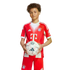 A young boy with curly hair wears a red and white FC Bayern Munich soccer uniform while holding a soccer ball against a plain white background.