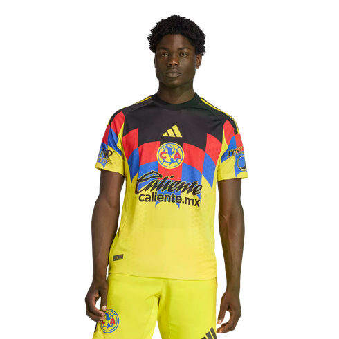 A man wearing a yellow Club América soccer kit with red and blue geometric patterns on the chest and sponsor logos, set against a black background.