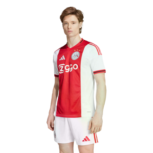 A man models the Ajax home kit: a red and white jersey with the "Ziggo" logo and white shorts, both featuring red Adidas stripes.