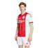 A man models the Ajax home kit: a red and white jersey with the "Ziggo" logo and white shorts, both featuring red Adidas stripes.