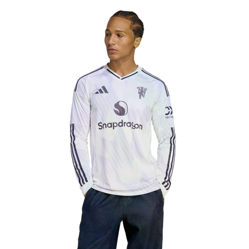 A man wearing a white long-sleeve Manchester United jersey with a Snapdragon logo, looking to the side against a black background.