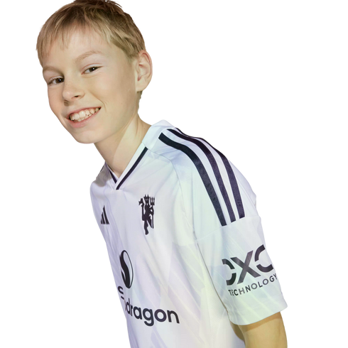 A young person with blonde hair smiles while leaning forward, wearing a white soccer jersey featuring several logos. The image is set against a solid black background.