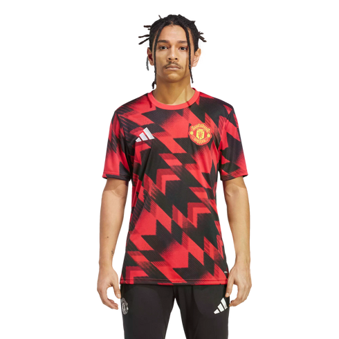 A man models a Manchester United training jersey featuring a red and black geometric pattern, paired with black Adidas joggers, against a plain white background.