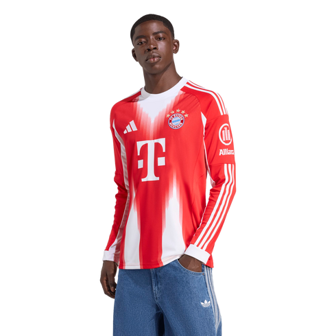 A Black man wearing a long-sleeved red and white Adidas FC Bayern Munich soccer jersey and blue jeans, standing with one hand in his pocket against a white background.