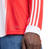 Close-up of a person wearing a red and white long-sleeved shirt with three white stripes on the sleeve, "AEROREADY" branding on the side, and blue denim jeans.