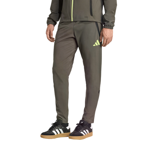 Lower body of a person wearing olive green Adidas track pants and black sneakers with white stripes and gum soles against a black background.