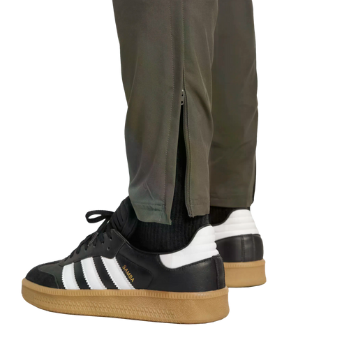 A low-angle view of black Adidas Samba sneakers with gum soles, worn with olive green pants featuring ankle zippers, set against a black background.