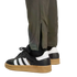 A low-angle view of black Adidas Samba sneakers with gum soles, worn with olive green pants featuring ankle zippers, set against a black background.