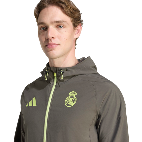 A man wearing an olive green Real Madrid Adidas windbreaker with neon yellow accents, posed against a black background.