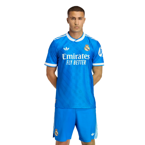 A man posing in a bright blue Real Madrid soccer kit with white Adidas and Emirates logos, standing against a solid black background.
